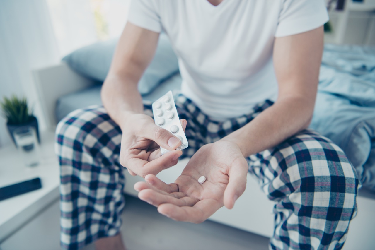 Cropped close up photo of sick man feel dizzy want use pills vitamins from virus cold high temperature sit on bed in morning house indoors wearing plaid checkered pajama
